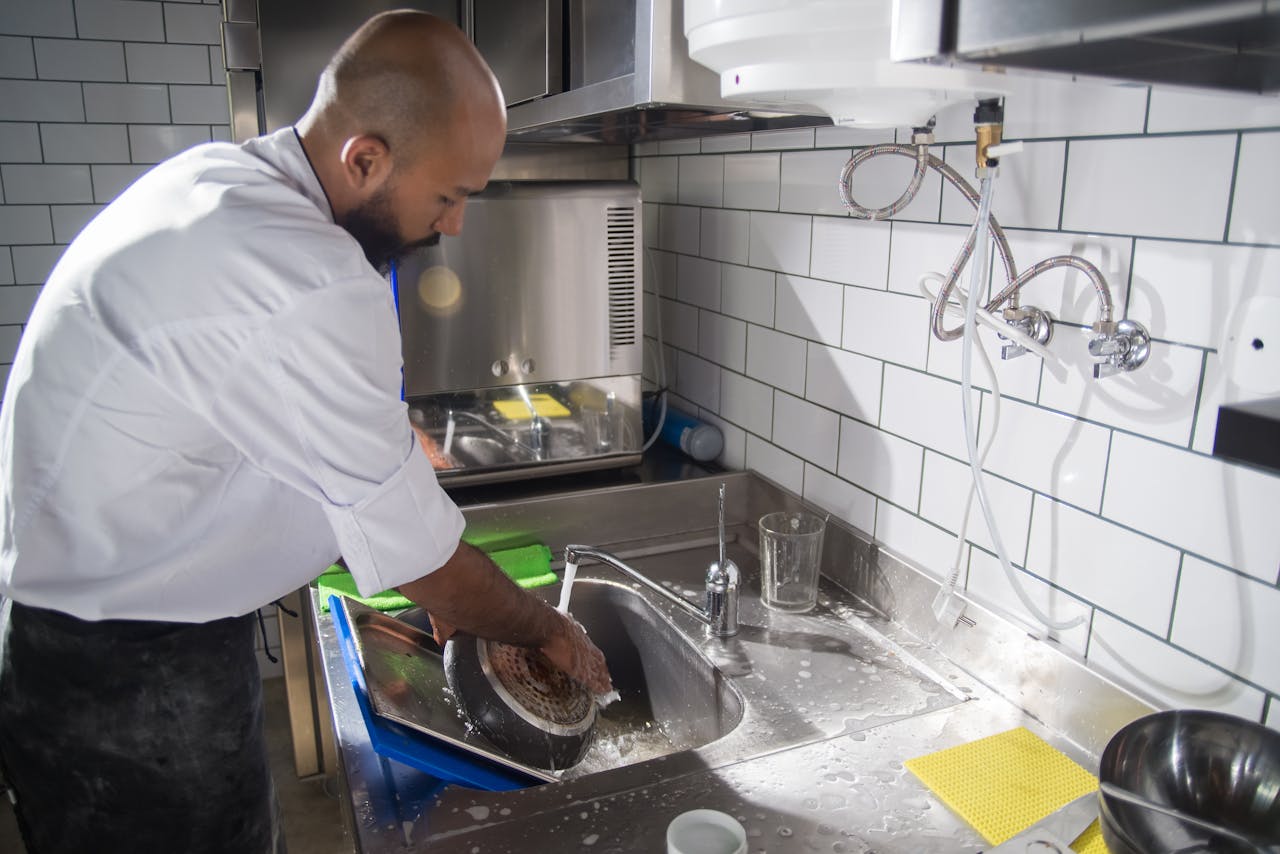 services-04 A chef cleans kitchen utensils in a stainless steel sink, maintaining hygiene standards.